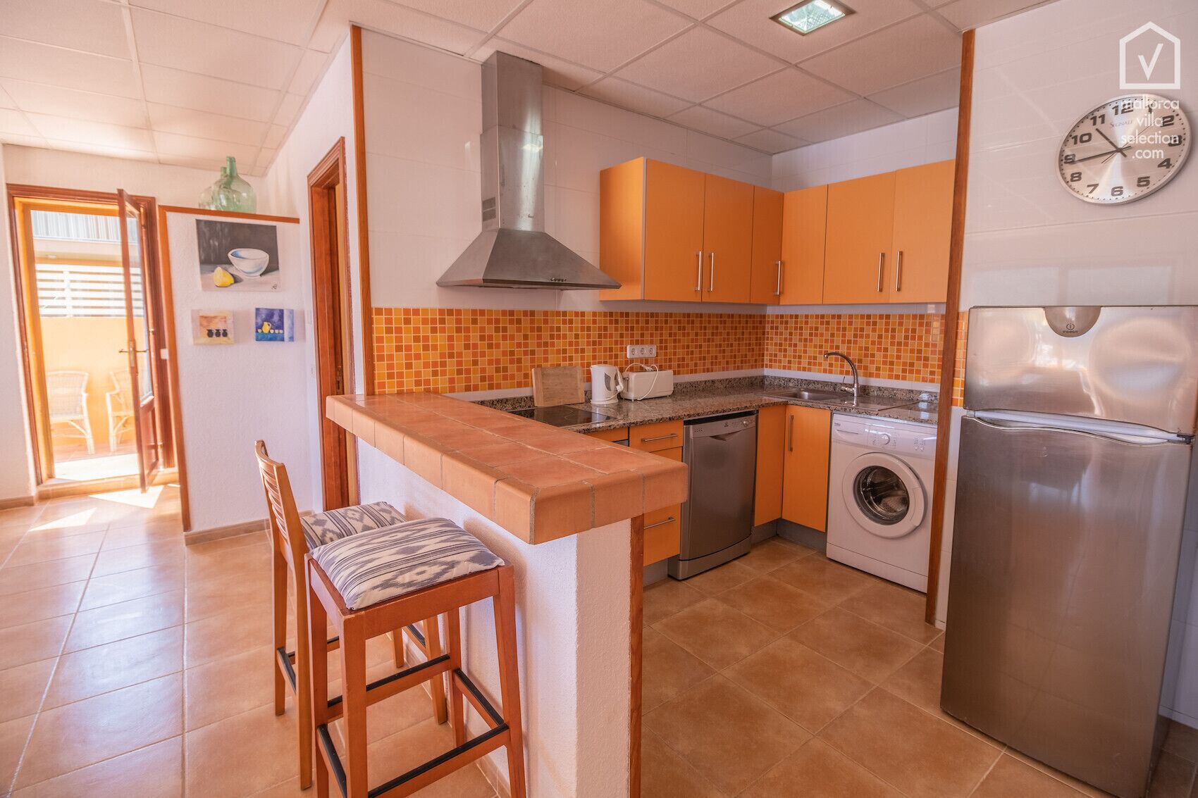 A compact kitchen with orange cabinets, a stainless steel range hood, a dishwasher, a washing machine, and a refrigerator.