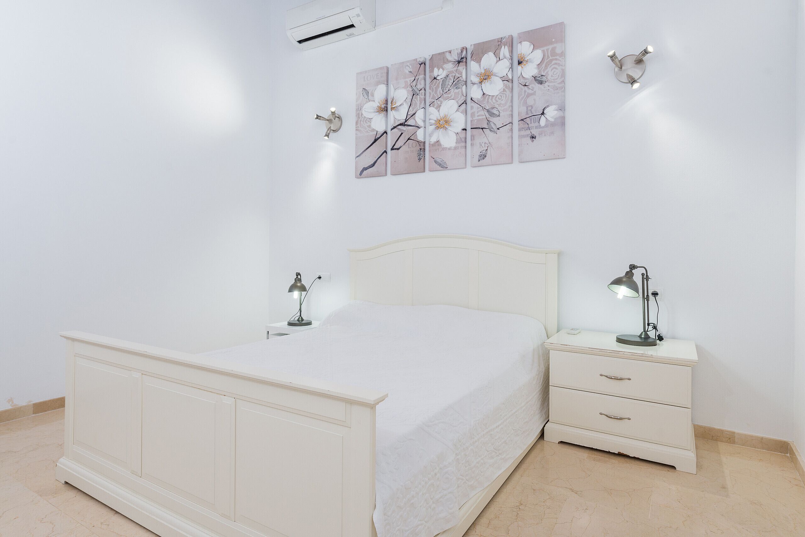 Ein Schlafzimmer mit einem weißen Bett, Nachttischen mit Lampen und einer Wand-Klimaanlage.
