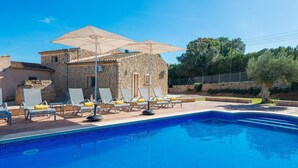 Ein Swimmingpool mit Liegestühlen und Sonnenschirmen, umgeben von Steinbauten und viel Grün.