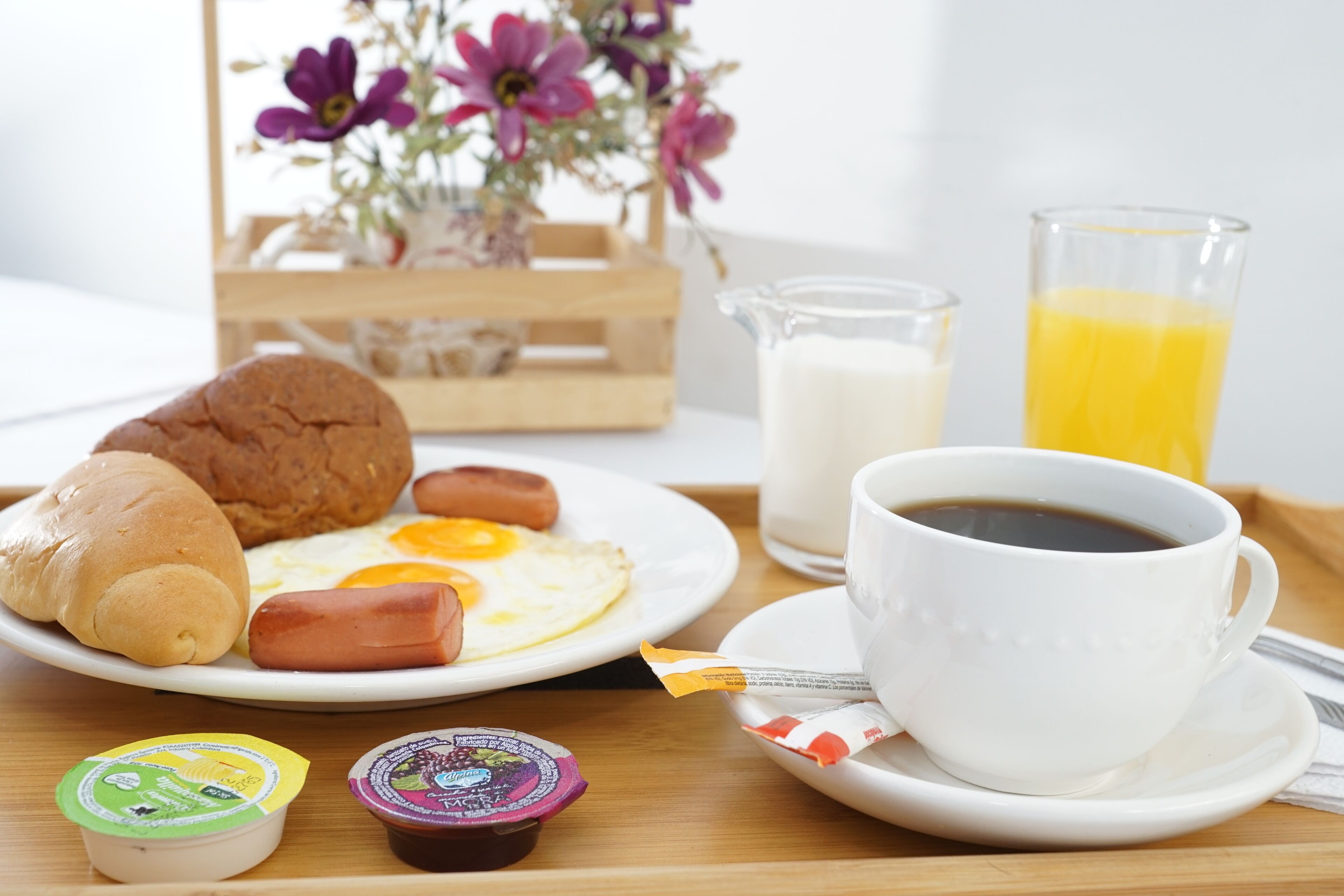 En frukostbricka med en croissant, en korv, ett stekt ägg och två burkar med pålägg, en kopp kaffe, ett glas apelsinjuice och en mjölkkanna.