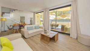 A modern living room with a sofa, coffee table, and dining area. It features a staircase, large windows, and a view of the outside. - Villa Jade II (Can Picafort)