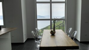 Et spiseområde med et træbord og stole, store vinduer med udsigt over et bakket landskab.