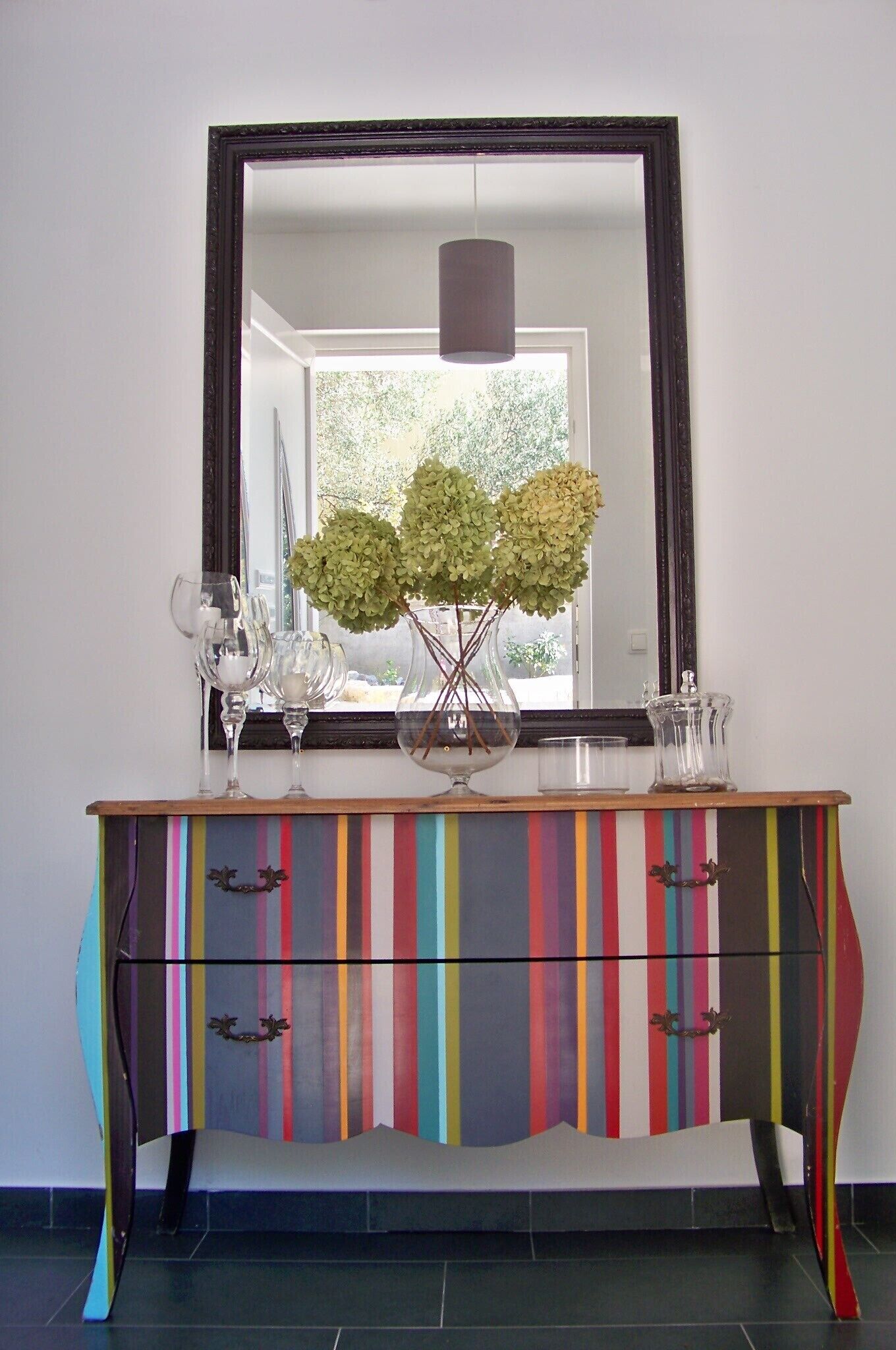 Ein bunter, gestreifter Schrank mit einem Spiegel darüber, eine Vase mit grünen Blumen und zwei Weingläsern.