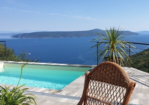 Una piscina con vistas al mar y las montañas, una silla de mimbre y una planta de palma.