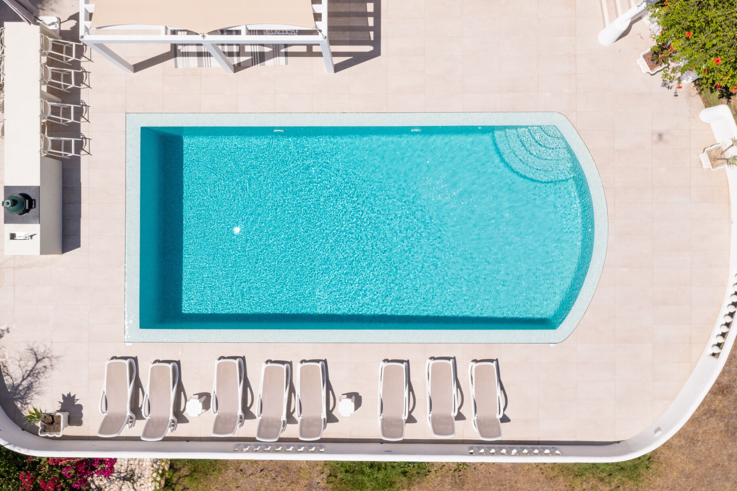 Vista aérea de una piscina rectangular con cubierta de baldosas, rodeada de sillones de descanso y una pequeña mesa.