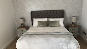 Una cama bien hecha con cabecera gris y dos mesitas de noche con lámparas.