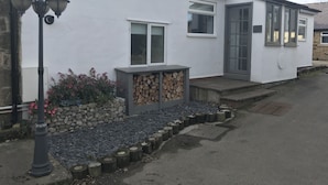 Un edificio blanco con un ventanal grande y una puerta, un pequeño porche con escalones, y una pila de leña.