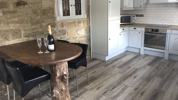 Un comedor con mesa y sillas de madera, una botella de champán y dos copas. Se observa una pared de piedra detrás de la mesa y una cocina con armarios y electrodomésticos blancos.