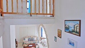 A room with a wooden balcony, a white door, a floral sofa, and a painting of a coastal scene. - Villa Mandola Rosa: Elegantly decorated, sea views, pool, A/C (Prodromi Latchi)