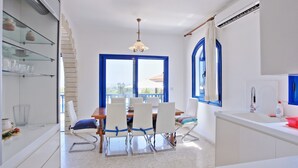 A modern kitchen with white cabinets, a dining area with a table and chairs, and a large window with blue frames. - Villa Mandola Rosa: Elegantly decorated, sea views, pool, A/C (Prodromi Latchi)
