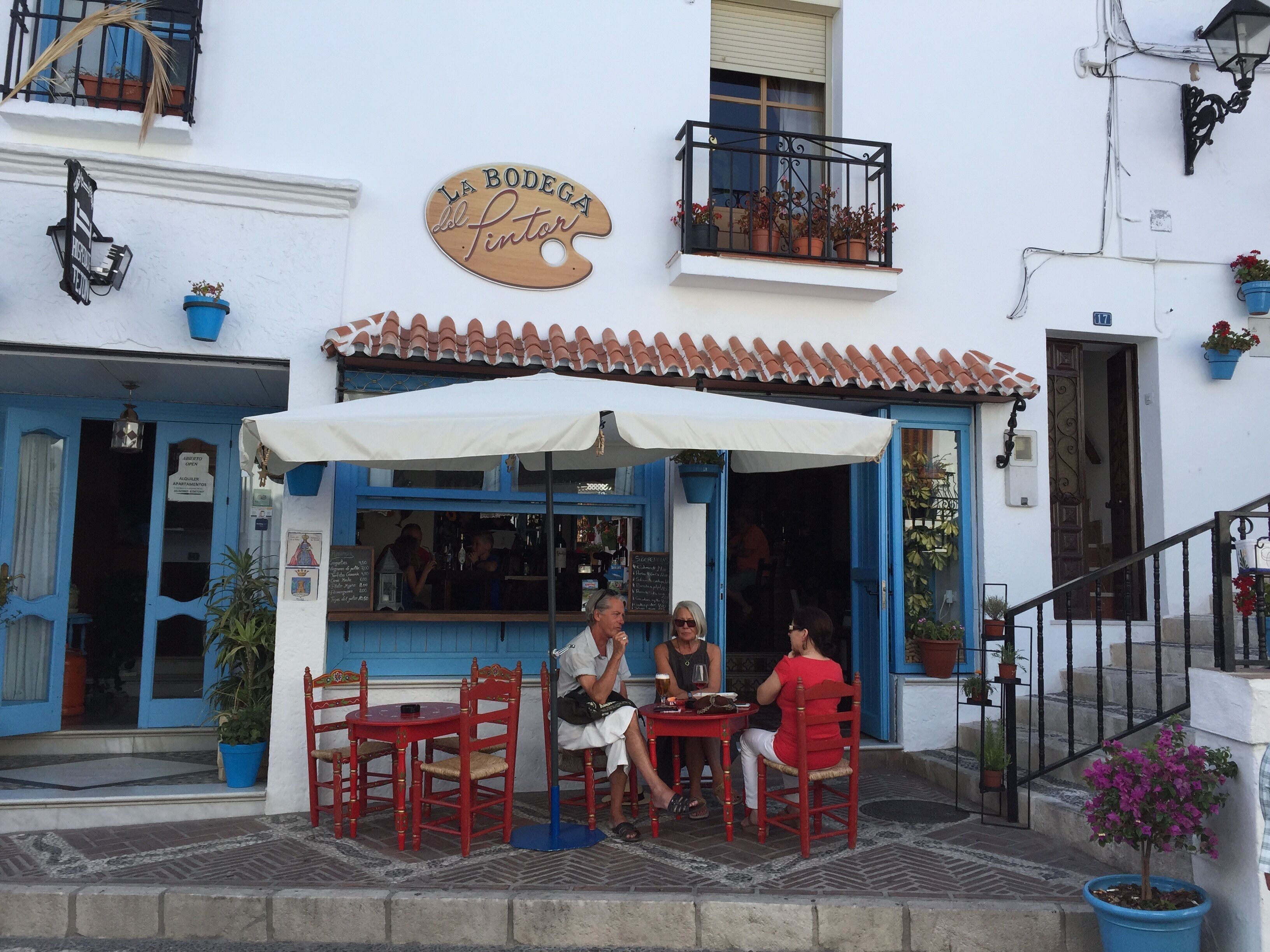 An outdoor café with red chairs and a white umbrella, a sign reading "La Bodega del Pintor", and a staircase leading to a door.
