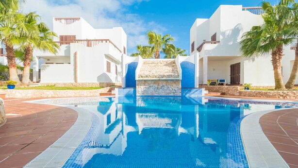 A swimming pool with a stone waterfall, surrounded by white buildings and palm trees. - MARINA D'OR IV CASA 5 - Chalet with shared pool in Cala D'Or. Free WiFi (Santanyí)