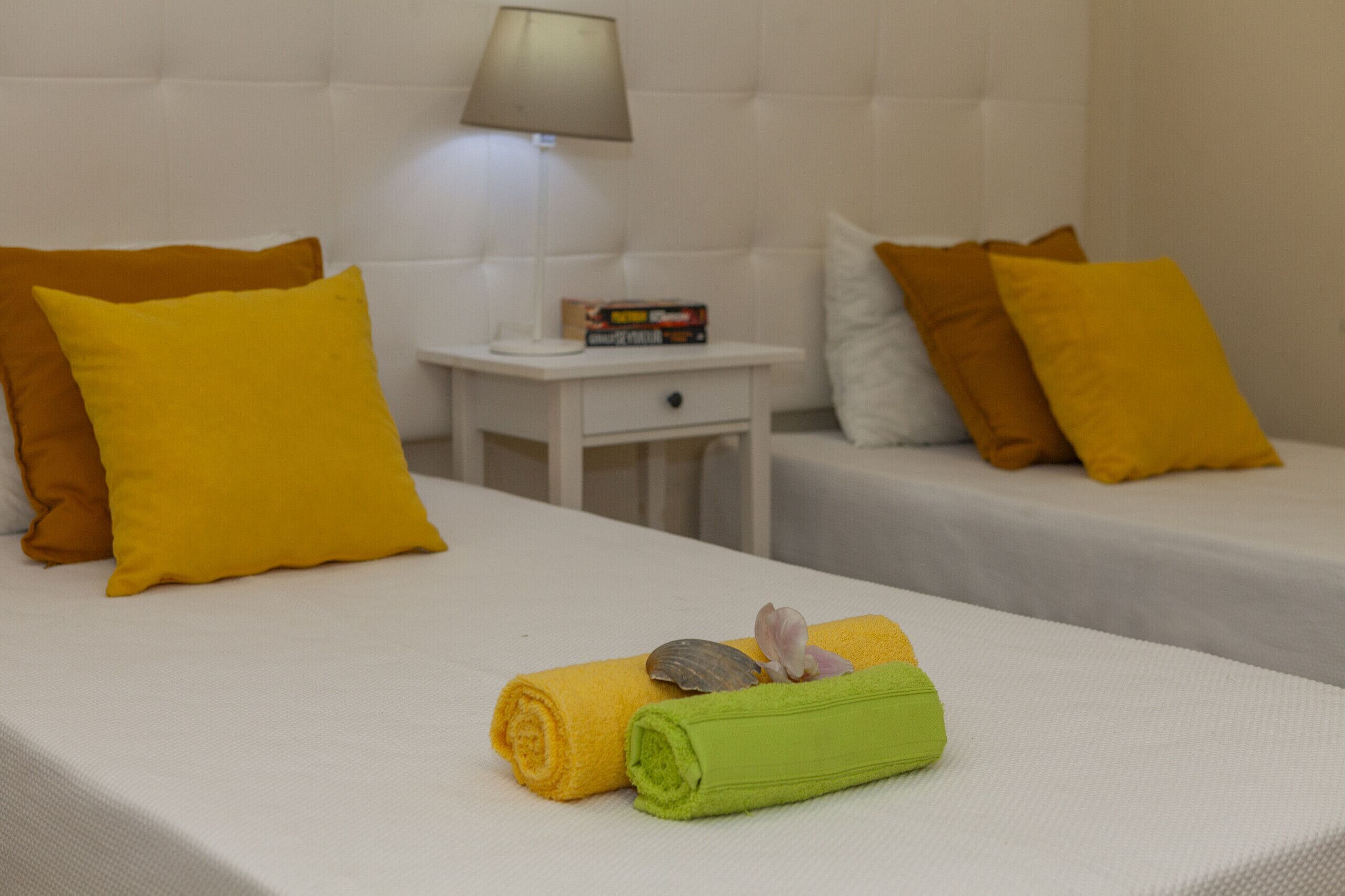 Una camera d'albergo con due letti, ognuno con cuscini gialli e marroni, un comodino con una lampada e un tavolino con dei libri.