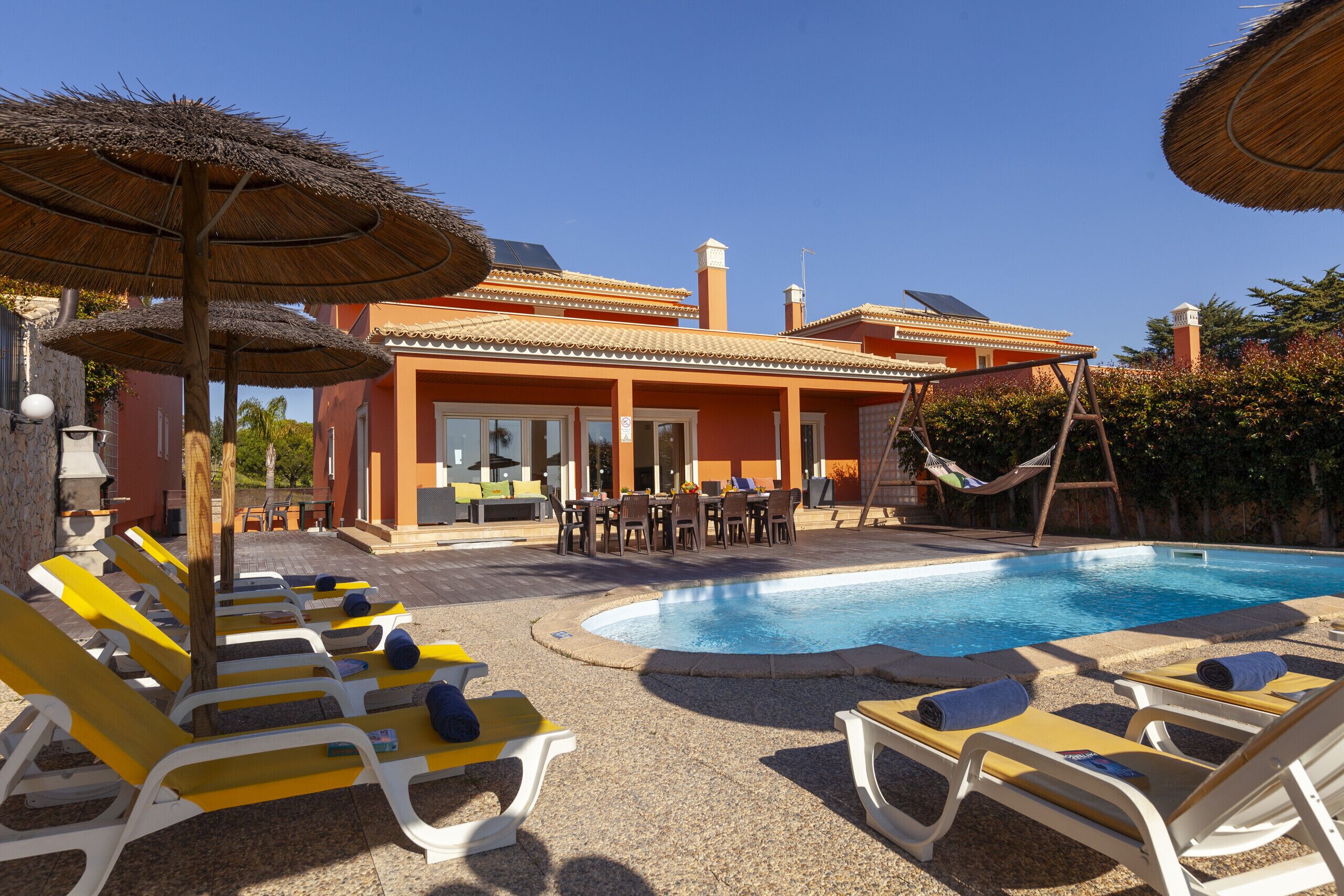 A pool area with sun loungers and umbrellas, a dining table with chairs, and a building with a tiled roof.