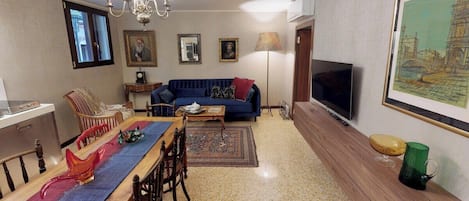 A living room with a sofa, television, dining table, chairs, and a chandelier.