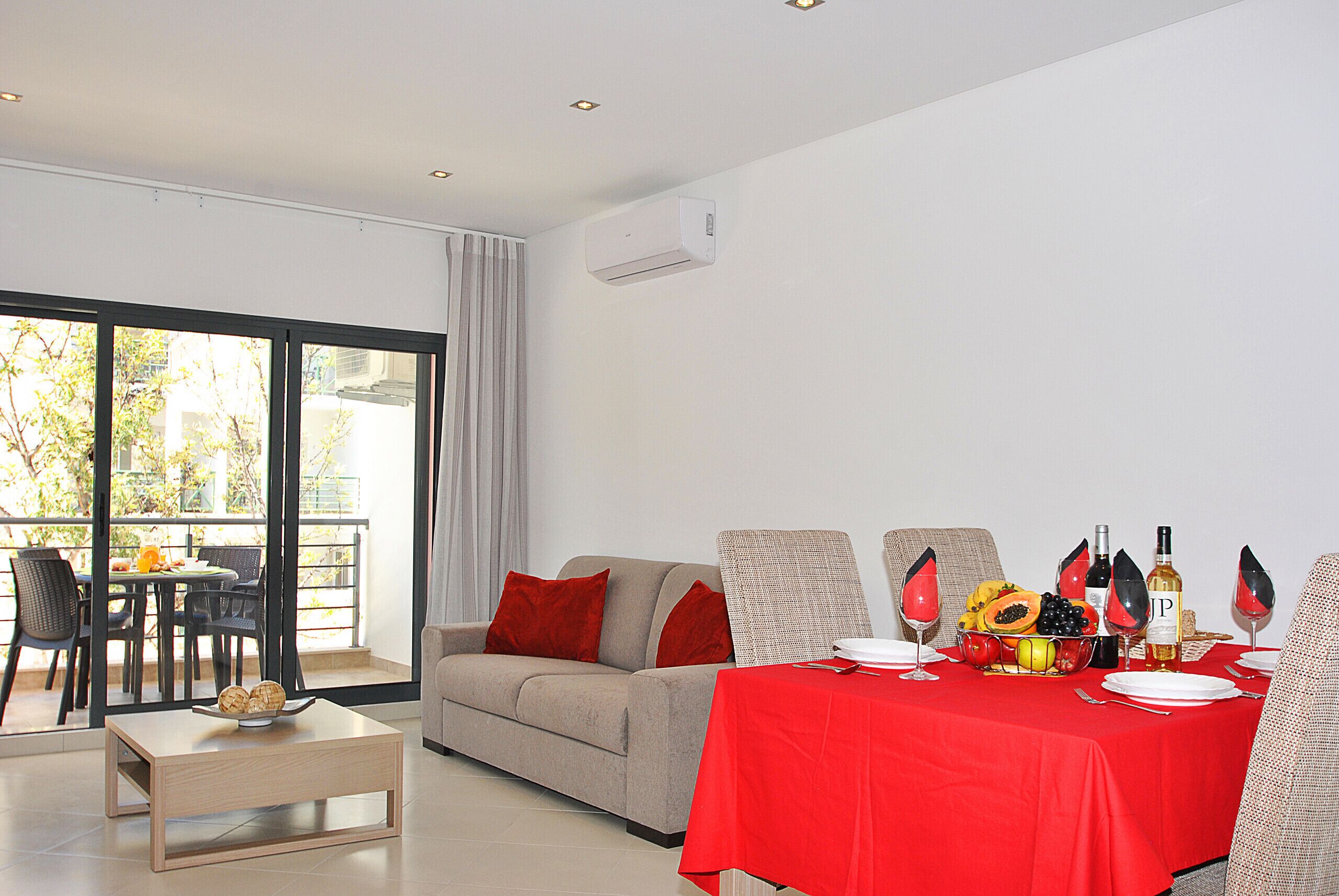 A modern living room with a dining area, a sofa, a table set for a meal, and a view through large windows.