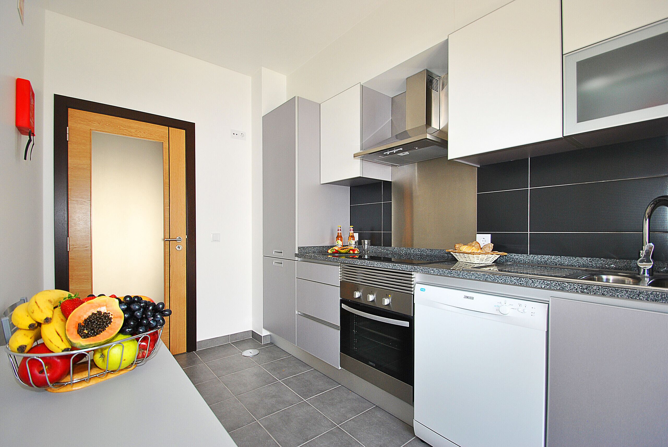 A modern kitchen with grey cabinets, a stainless steel range hood, a dishwasher, and a fruit bowl on the counter.