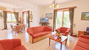 Un salon comprenant un canapé, des fauteuils, une table basse et un téléviseur. On y trouve également une table à manger avec des chaises et un lustre.