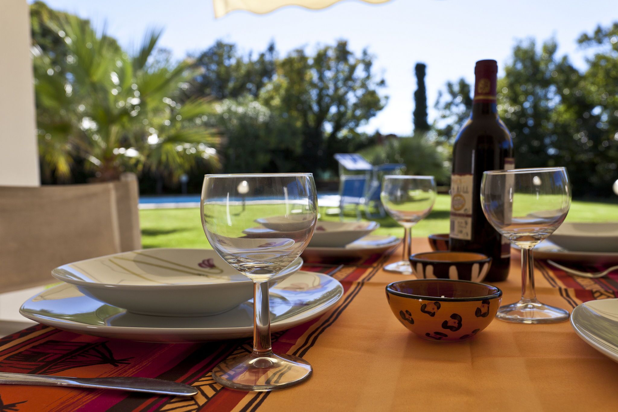 Un tavolo apparecchiato all'aperto, con piatti, ciotole e bicchieri da vino, una bottiglia di vino e vista su un giardino con palme.