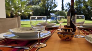 Een buitentafel gedekt met borden, kommen en wijnglazen, een fles wijn, en een uitzicht op een tuin met palmboompjes.