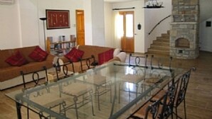 Una sala de estar con una mesa de centro de vidrio, un sofá marrón con cojines rojos, una chimenea de piedra y una escalera que sube a otro nivel.