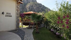 Una villa llamada "VIA JAMARA" con un sendero de piedra que conduce a un área de estar cubierta, rodeada de exuberante vegetación y un telón de fondo montañoso.