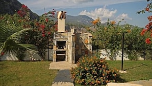 Un edificio de piedra con chimenea, rodeado de un jardín con plantas en flor y una montaña al fondo.