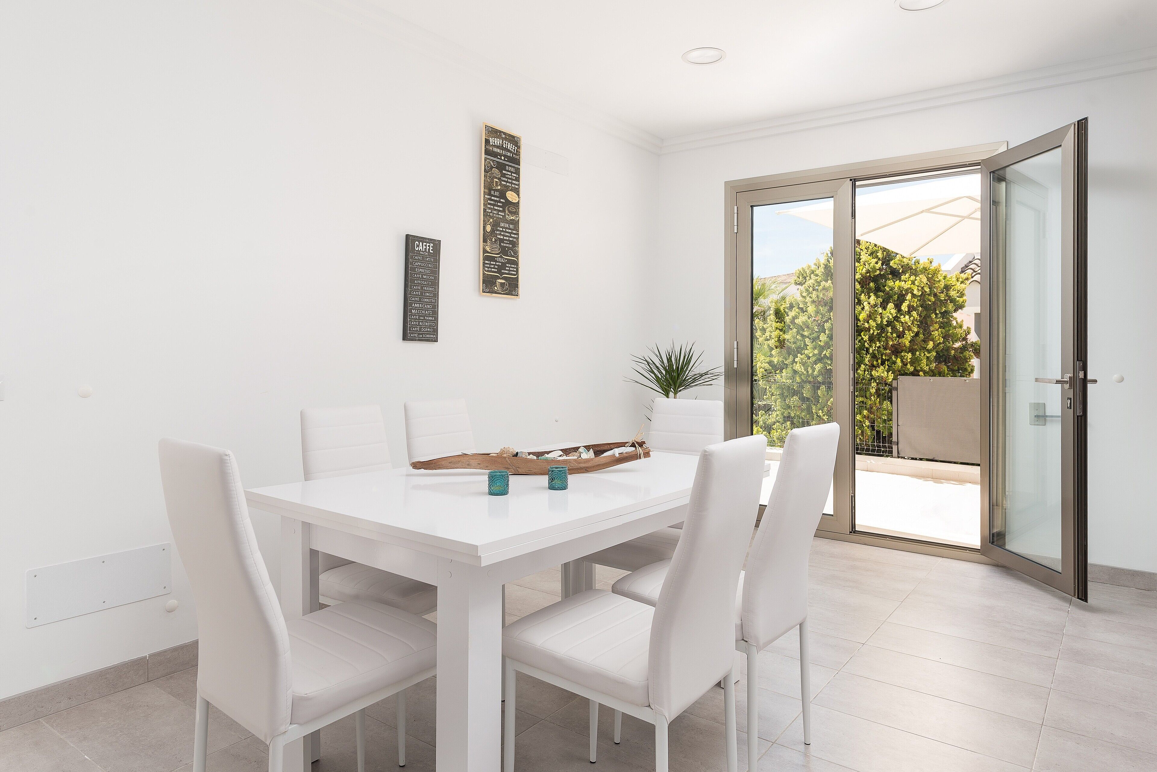 Un coin repas avec une table et des chaises blanches, une plante et une porte-fenêtre coulissante donnant sur un espace extérieur.