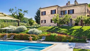 Ein Schwimmbecken mit Liegestühlen, ein Steinhaus mit zahlreichen Fenstern und ein gepflegter Garten.