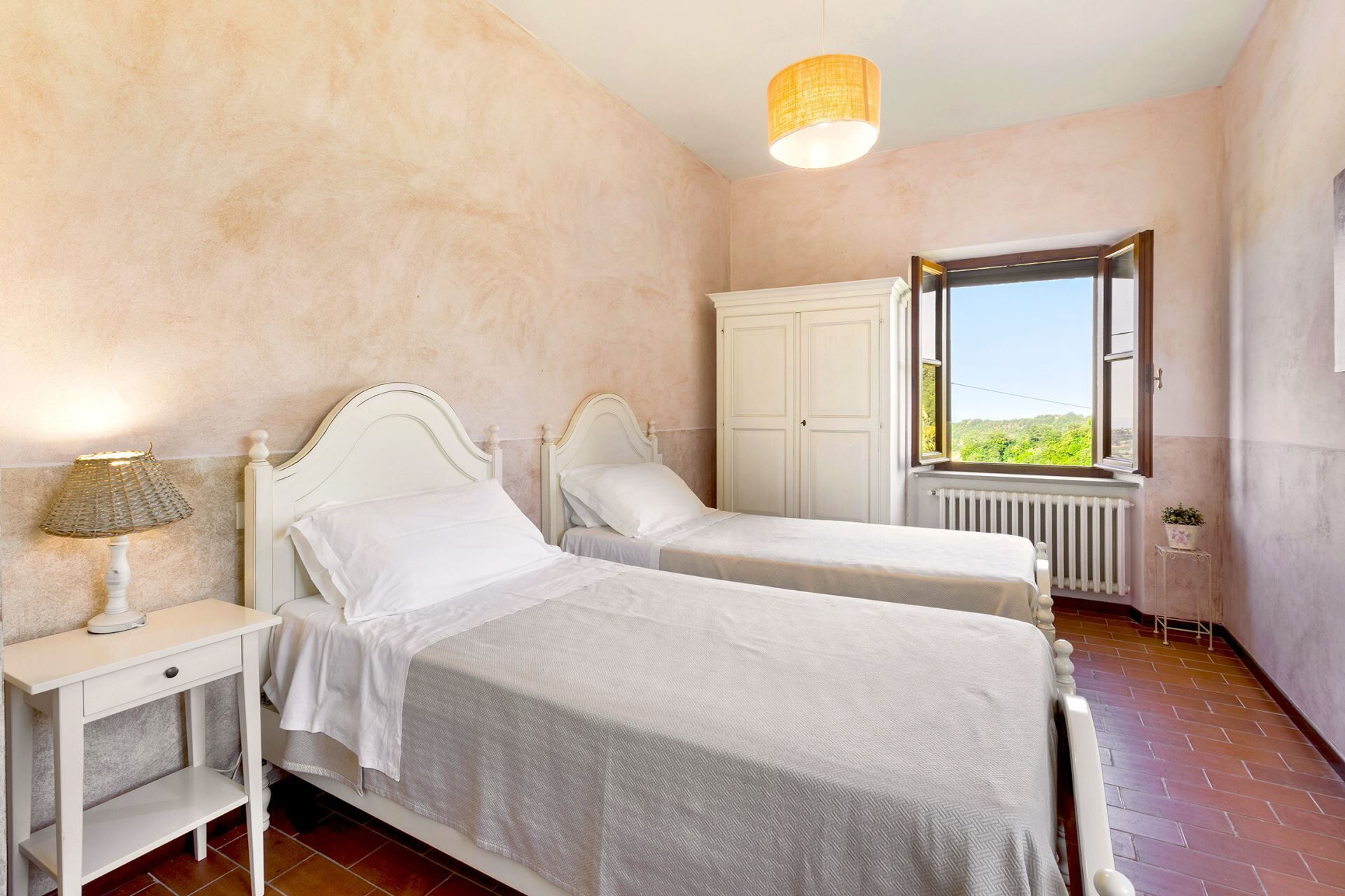 Een slaapkamer met twee eenpersoonsbedden, een nachtkastje met lamp, een kledingkast en een raam met uitzicht op groen.