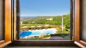 Blick auf ein Schwimmbecken mit Liegestühlen und Sonnenschirmen, umgeben von Grünflächen und Hügeln.
