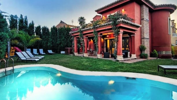 Una piscina con lettini, un edificio con colonne rosse e un prato curato.