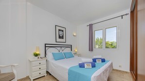 A bedroom with a bed, bedside tables, a nightstand, a chair, and a window with curtains. - Villa Heura (Alcúdia)