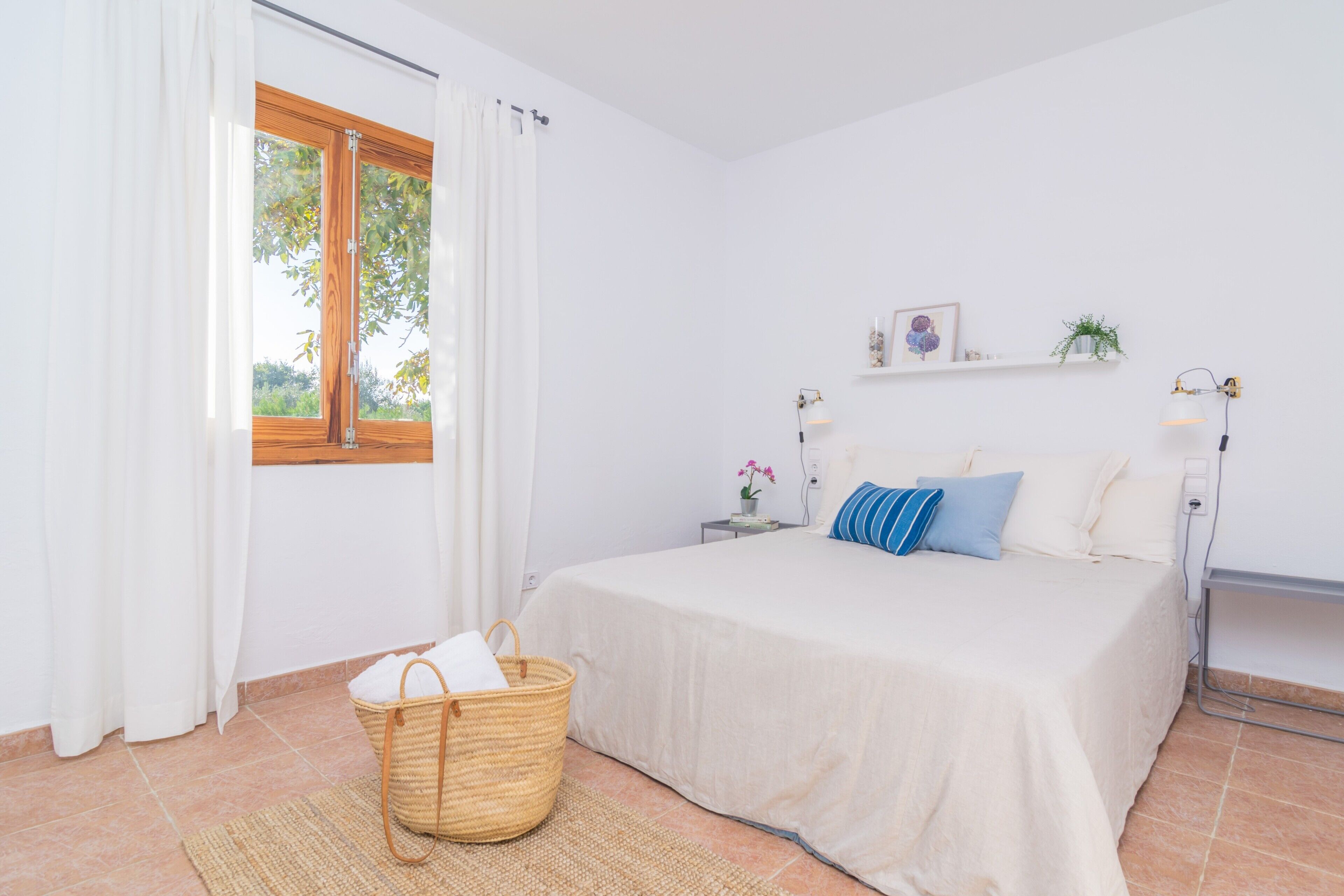 A bedroom with a bed, a bedside table, a lamp, a window with curtains, a wall shelf with decorative items, and a wicker basket on the floor.