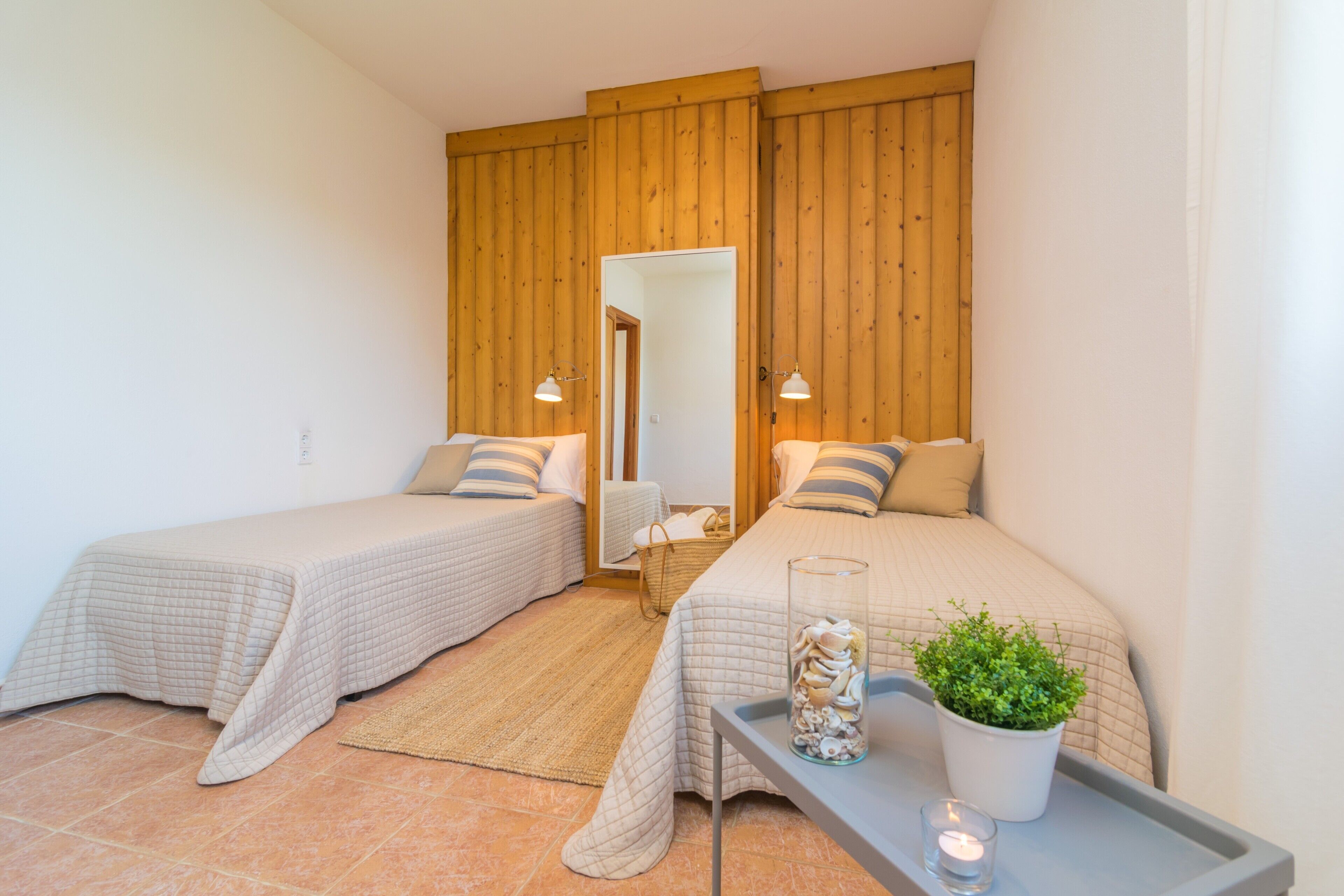 A bedroom with two beds, a wooden headboard, a mirror, a bedside table with a lamp, a small plant, and a glass jar with seashells.