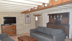 Een woonkamer met een stenen muur, een houten kast met flessen, een grijze bank, een houten salontafel en een televisie op een houten dressoir.