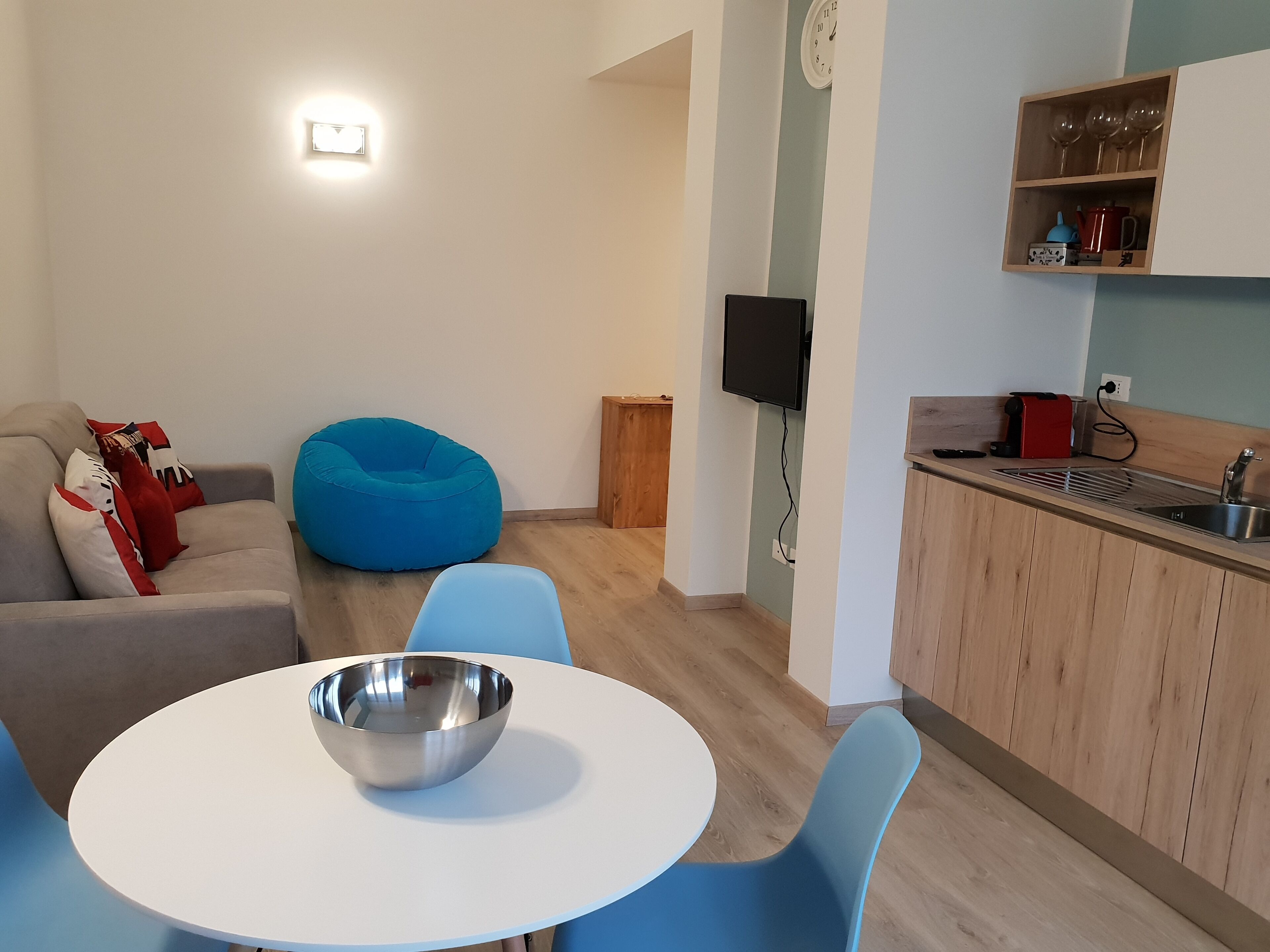 A modern living room with a sofa, blue bean bag, and a dining table with a metal bowl. A kitchen area with wooden cabinets and a TV is visible in the background.