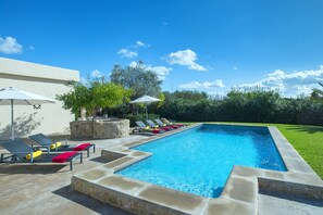 A rectangular swimming pool with a stone border, surrounded by lounge chairs and umbrellas, set against a backdrop of a clear blue sky and greenery.