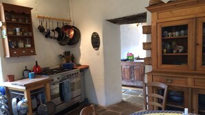 Eine Küche mit Herd, Backofen und hängenden Töpfen. Ein Tisch mit Stühlen und einer gemusterten Tischdecke steht ebenfalls dort.
