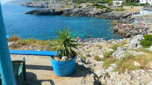 Une vue sur la côte, avec une plage rocheuse, une mer bleue et limpide, et quelques personnes qui profitent de l’eau.