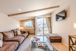 A modern living room with a sofa, ottoman, and glass coffee table, a TV mounted on the wall, and a view of a mountainous landscape through large windows. - Luxurious penthouse 3 bed in the center (MORZINE)