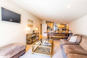 A modern living room with a sofa, coffee table, and a TV. There is a kitchen in the background with wooden cabinets and a microwave. - Luxurious penthouse 3 bed in the center (MORZINE)