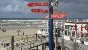 Une jetée au bord de la plage, agrémentée de panneaux indicateurs, une tour de surveillance pour les sauveteurs et une plage de sable où se trouvent des gens.