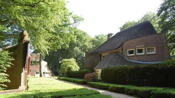 Un bâtiment traditionnel en briques, doté d’un toit de chaume, entouré de haies et d’arbres bien entretenus.