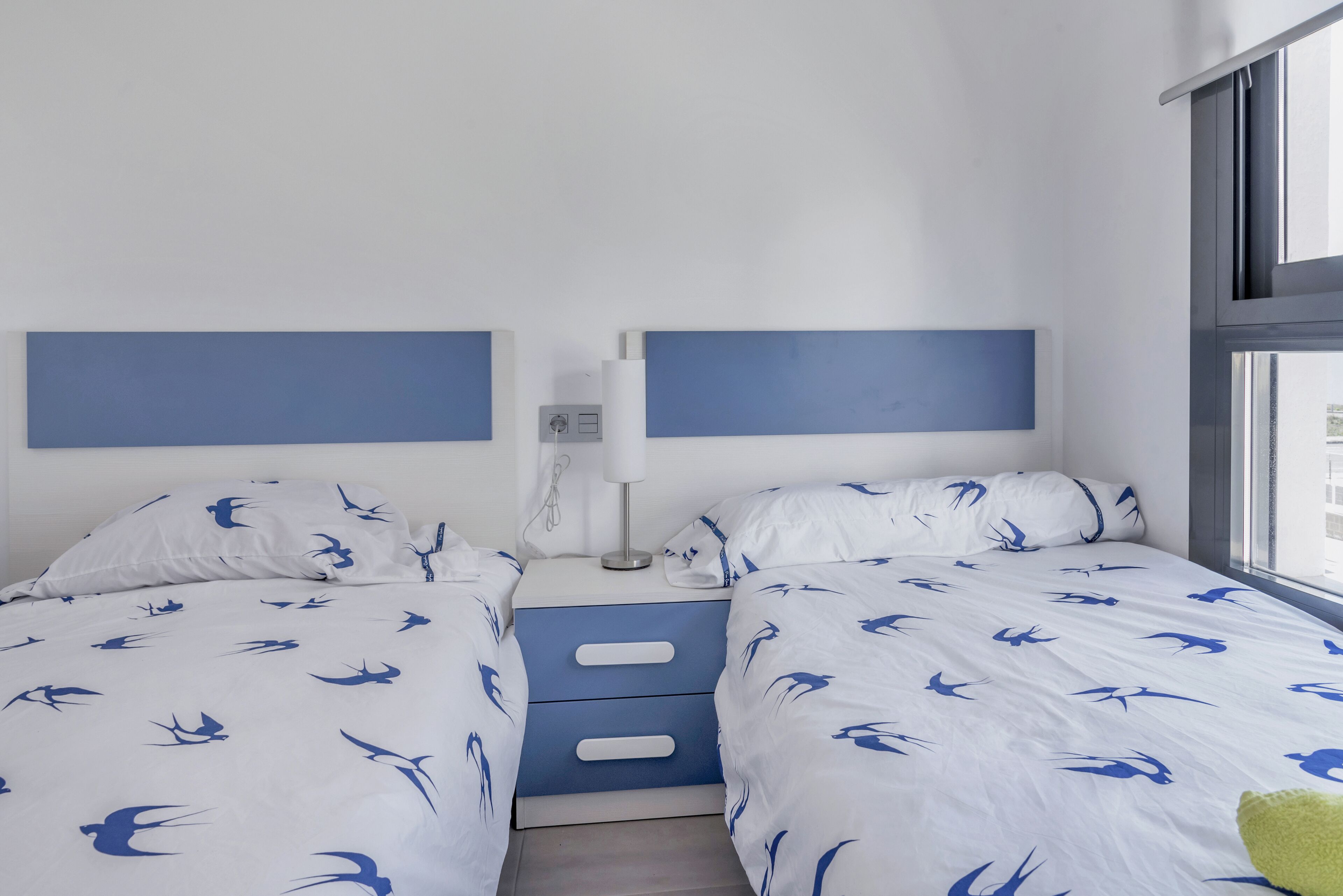 A twin bedroom with two beds, each with a white and blue bird pattern bedspread, a bedside table with a lamp, and a window with a view of the outside.