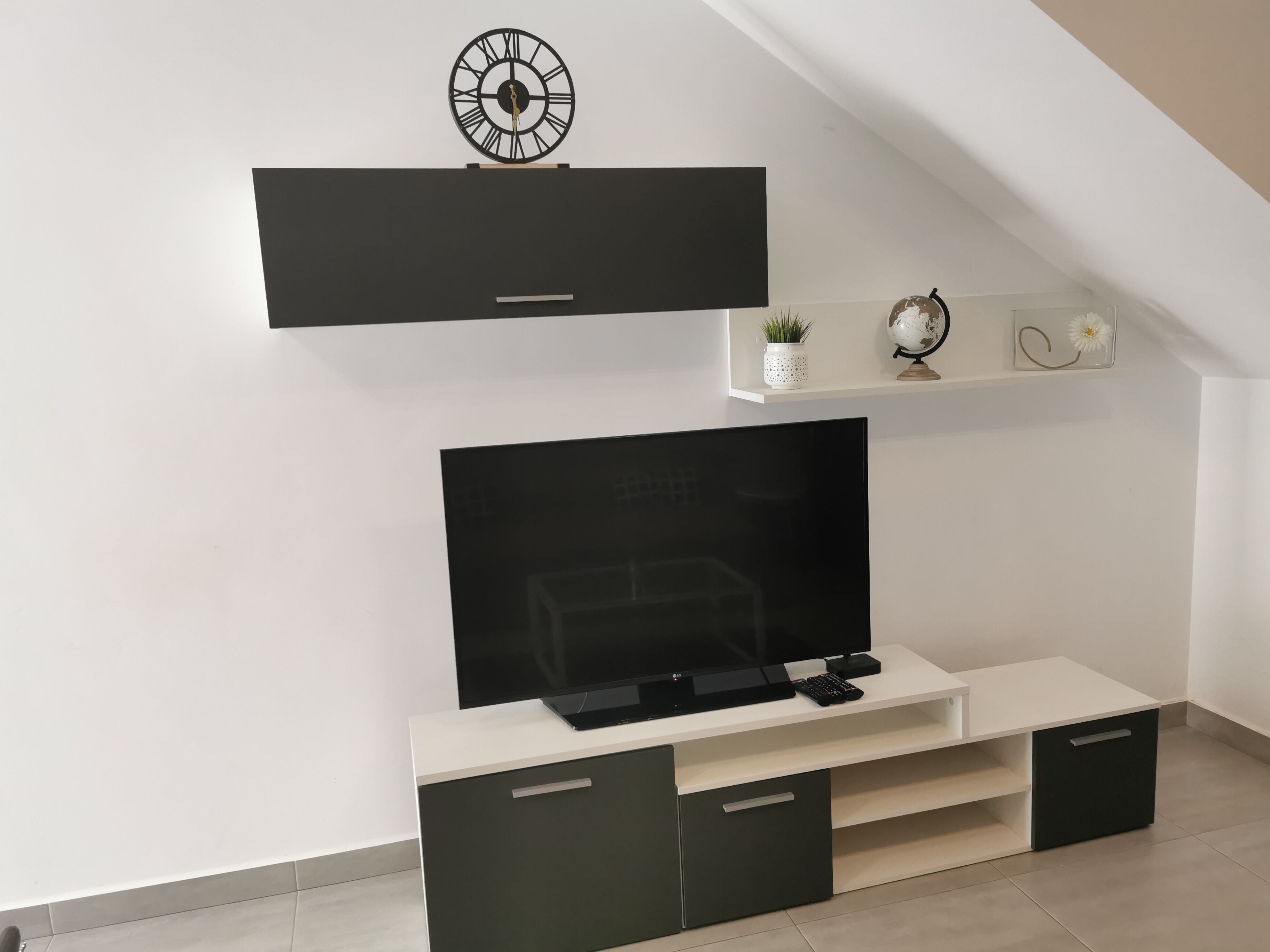 A modern living room with a flat-screen TV mounted on a white console, a black entertainment unit above, and a small shelf with decorative items.