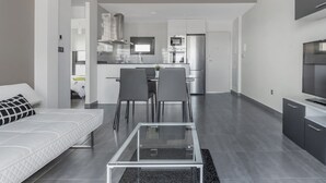 Una sala de estar moderna con una mesa de centro de vidrio, un sofá y un comedor con sillas, con una cocina al fondo.