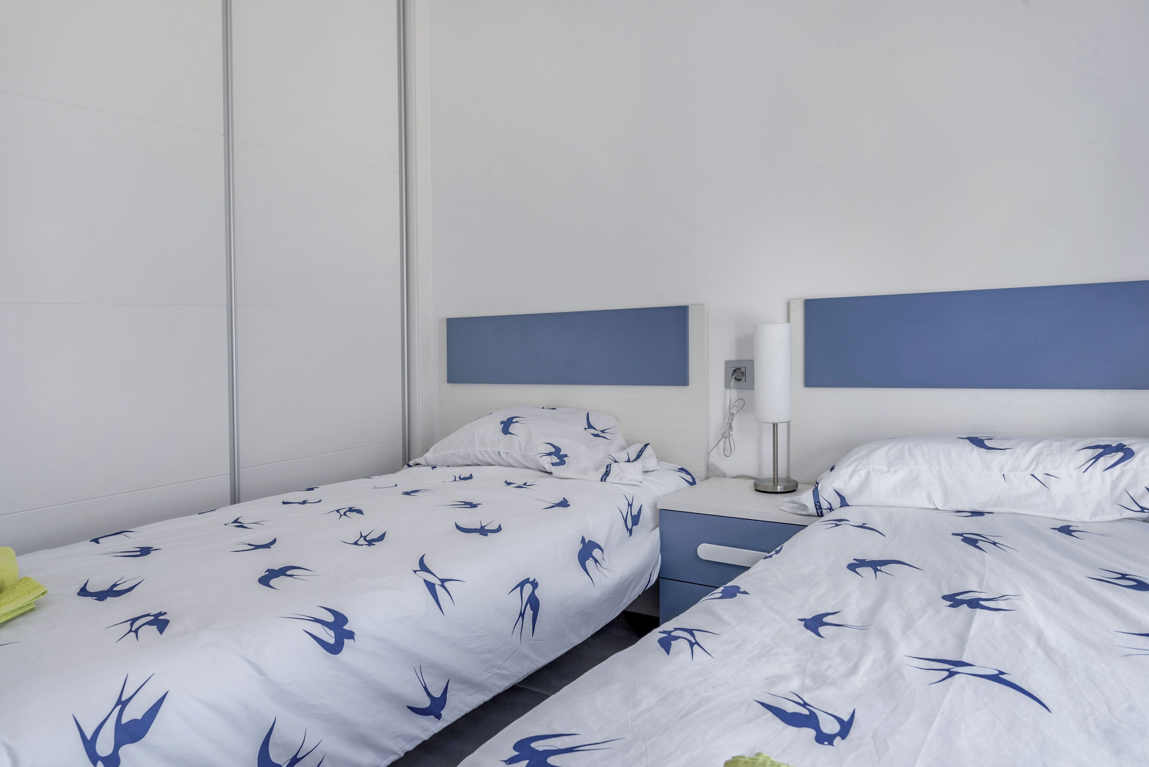 Une chambre d’hôtel avec deux lits simples, chacun doté d’une tête de lit bleue et de draps blancs ornés d’un motif représentant des oiseaux. On y trouve une table de chevet avec une lampe et un téléphone.