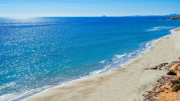 Una playa con agua azul cristalina, orilla arenosa y montañas a lo lejos.