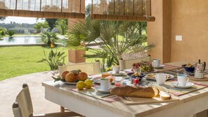 Ein Frühstückstisch mit Früchten, Brot und Getränken, mit Blick auf einen Pool und Grünflächen.